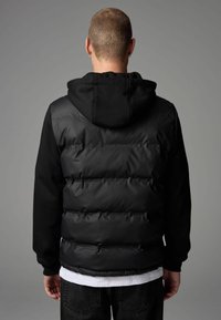 Gilet à capuche noir en nylon rembourré avec un tissu de pull contrastant, dotée d'un dos texturé et de poignets côtelés. Design simple et monochrome.
