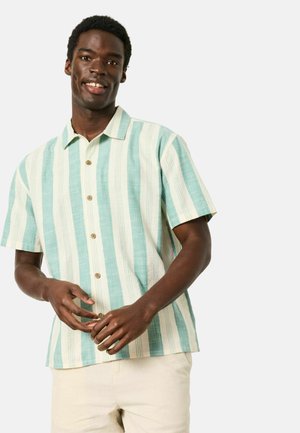 Mann lächelnd, trägt ein kurzärmeliges Hemd mit senkrechten grünen und weißen Streifen und hellen Hosen, die Hände vor dem Körper verschränkt.