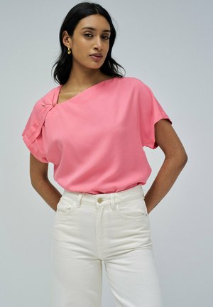 Top rosa de manga corta con un diseño drapeado y sutil recogido en el hombro, que presenta un detalle decorativo en dorado. Combinado con pantalones blancos.