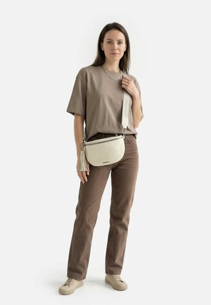 Mujer con pantalones marrones, camiseta oversize taupe, zapatillas beige y bolso blanco cruzado con flecos, de pie contra un fondo blanco.
