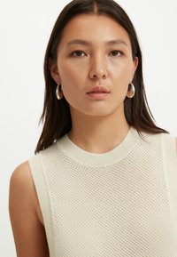 Ung kvinna med långt mörkt hår som bär silverfärgade ringörhängen och en ärmlös krämfärgad stickad topp, som tittar rakt fram med neutralt uttryck.