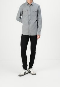 Camicia grigia con bottoni, dotata di due tasche sul petto, morbida al tatto, abbinata a jeans skinny neri e sneakers bianche con accenti grigi.