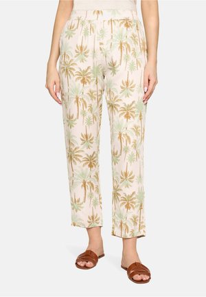 Woman wearing light-colored pants with green and brown palm tree print and brown slide sandals, standing with hands relaxed.