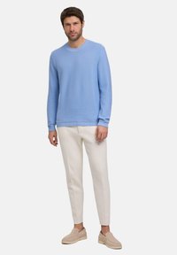 Lichtblauwe gebreide trui met geribde hals en manchetten, gecombineerd met witte smalle broek en beige instap schoenen. Effen achtergrond.