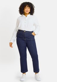 Vaqueros de denim oscuro de cintura alta con pierna recta, que presentan costuras doradas y bolsillos delanteros, combinados con una camisa blanca de botones.