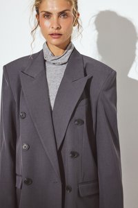 Blazer grigio doppiopetto con grandi bottoni neri, caratterizzato da rever appuntiti e una vestibilità comoda, indossato sopra un dolcevita grigio.