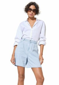 Light blue denim high-waisted shorts with front pleats and side pockets, paired with a white button-up shirt. Large dark sunglasses worn.