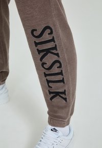 Bruna joggers med svart "SIKSILK" broderi längs sidan, med en texturerad tygkvalitet och muddar vid anklarna. I kombination med vita sneakers.