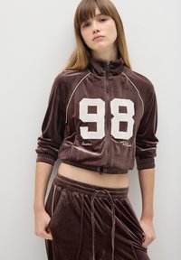 Giacca a track in velluto marrone con stampa bianca "98" e ricamo "Boston". Presenta una chiusura con zip, colletto alto e polsini elastici. Abbinata a pantaloni coordinati.