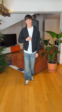 Schwarzer Cardigan über einem weißen T-Shirt, blaue Jeans mit weitem Bein und silberne Sneakers. Innenraum mit einer Pflanze und einem braunen Sofa.