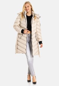 Beige Steppjacke mit gepolstertem Design, durchgehendem Reißverschluss, abnehmbarem Kapuzenhut und Strickbündchen, getragen über einem schwarzen Oberteil und hellblauen Jeans.