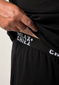 Mano levantando una camisa negra para revelar una cinturilla negra con el texto blanco "RELAX + CHILL" en pantalones o pantalones cortos negros.
