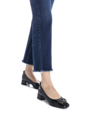 Zapatos de charol negro de tacón medio con punta redonda y un logotipo decorativo en la parte frontal, combinados con jeans oscuros de dobladillo deshilachado.
