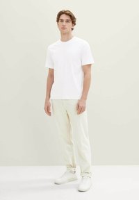 Maglietta bianca in cotone con scollo rotondo, abbinata a pantaloni beige chiaro a taglio affusolato e sneakers bianche, su sfondo neutro.