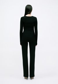 Top lungo a maniche ribattute nere con un design aderente, abbinato a pantaloni neri a vita alta, mostrato da dietro su uno sfondo chiaro e uniforme.