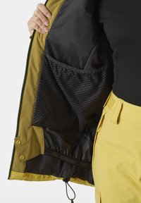Innenansicht einer grünen und gelben Jacke, die eine dunkle Netzinnentasche und einen elastischen Saum zeigt. Das Außenmaterial ist glatt und verfügt über Druckknöpfe und Kordelzüge.