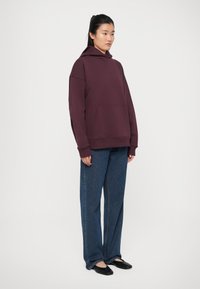 Hoodie oversize bordeaux con tasca frontale, polsini a costine e vestibilità comoda, abbinato a pantaloni in denim blu a gamba larga. Scarpe nere piatte.