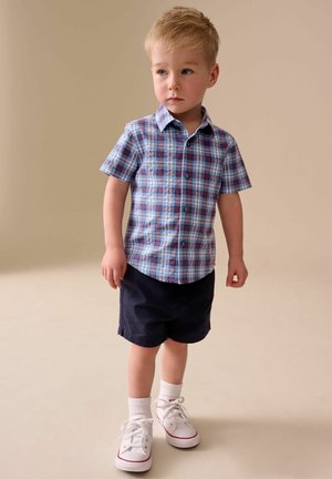 Băiat tânăr purtând o cămașă cu mânecă scurtă în carouri albastre, pantaloni scurți bleumarin, șosete albe și adidași albi, stând pe un fundal neutru.
