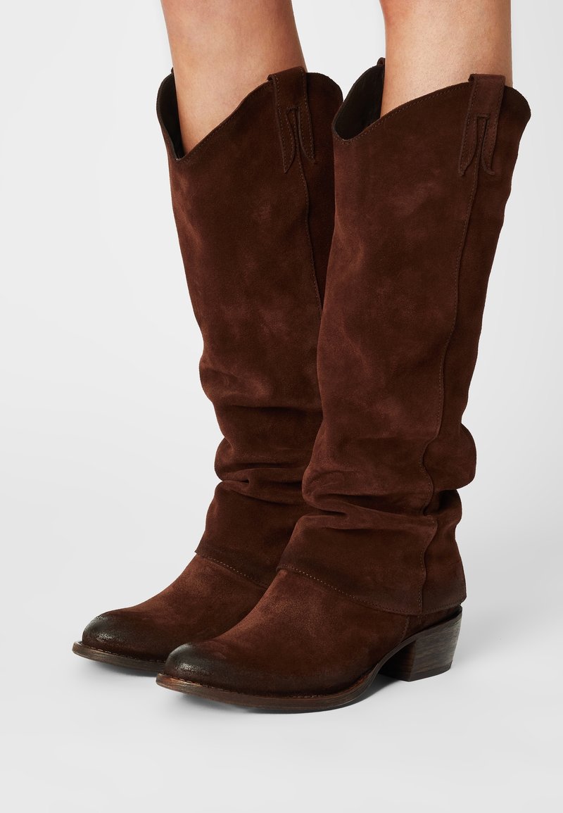 Bottes marron en velours côtelé, mi-mollet, avec des orteils ronds et de petits talons en bois empilés, portées sur des jambes nues.