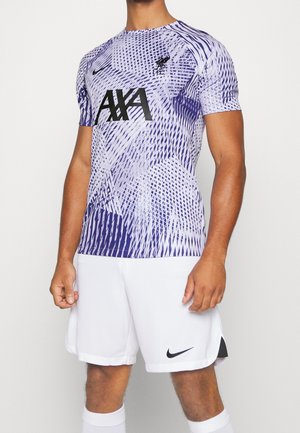 Homme portant un maillot de football de Liverpool violet à motifs géométriques avec le logo AXA, un short blanc Nike et des chaussettes blanches sur un fond uni.