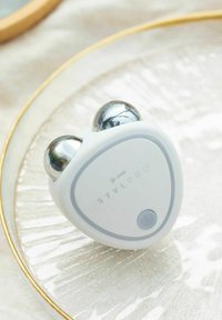 StylPro facial massager with a triangular white body, two silver balls on top, and a gray power button. Textured surface with slight shine.
