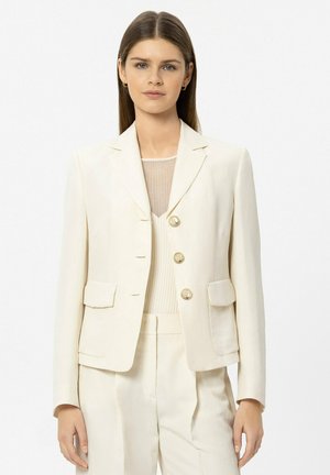 Frau mit glatten braunen Haaren, die ein cremefarbenes, maßgeschneidertes Blazer, passende hoch taillierte Hosen und ein geripptes Oberteil trägt.