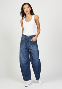 Leichtes weißes Tanktop kombiniert mit oversized blauen Jeans. Die Jeans verfügen über frontale Taschen und sichtbare Nähte. Weiße Sneakers runden das Outfit ab.