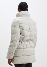 Manteau matelassé en gris clair avec un col haut. Présente des coutures en chevrons, une taille ceinturée et un matériau doux et isolant.