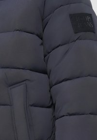 Kurtka puchowa w kolorze granatowym, wykonana z pikowanej tkaniny z wypełnionymi segmentami. Posiada kwadratową czarną metkę z napisem "JOOP! JEANS" na ramieniu.