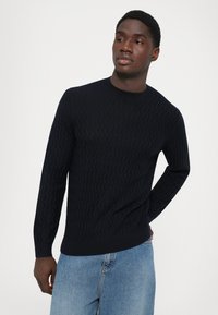 Mann iført en strukturt bukseblå genser og lyseblå jeans, og ser til siden med en hånd bak ryggen.