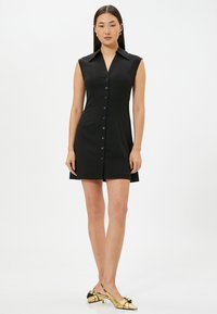 Sort ærmeløs kjole med krave, der har en frontlukning med knapper og en lige skæring. Blankguld slingback-sko med en sløjfe.