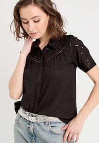 Chemise noire à manches courtes avec des motifs floraux brodés et un col boutonné, associée à un jean taille haute bleu clair et une ceinture en argent.