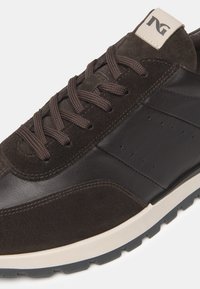 Sneaker marrone con una combinazione di camoscio e pelle. Lacci marrone scuro con perforazioni sui lati, suola bianca e un piccolo marchio.