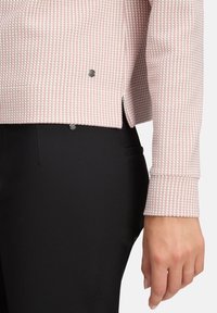 Nahaufnahme einer Hand, die schwarze Hosen und ein langärmliges Oberteil in Pink und Weiß mit strukturiertem Design, seitlichen Knopfdetails und einem kleinen Schlitz trägt.