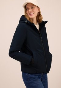 Giacca a vento blu scuro con cappuccio e zip, dotata di tasche laterali e una texture liscia. Indossata sopra abbigliamento di colori chiari e jeans.