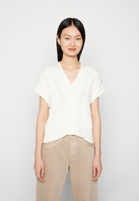 Weißer Strickcardigan mit Zopfmuster, kurzen Ärmeln und V-Ausschnitt mit Knöpfen auf der Vorderseite. Getragen über beigen Hosen.