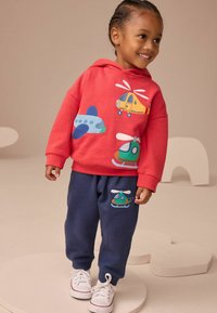 Bambino sorridente con felpa rossa con toppe di aerei e elicotteri, pantaloni blu navy con toppa di elicottero e sneakers bianche, in piedi su uno sfondo chiaro.