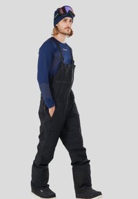 Czarne kombinezony narciarskie z regulowanymi ramiączkami, wieloma zamykanymi kieszeniami, noszone na niebieskiej koszuli z długim rękawem, w połączeniu z czarno-białymi butami śniegowymi.
