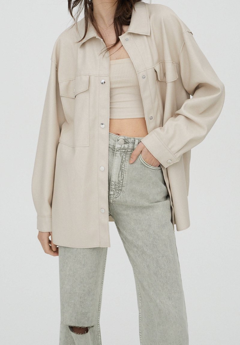 Femme portant une chemise beige oversize boutonnée, un crop top côtelé beige, et un jean gris clair taille haute déchiré au genou, main dans la poche.