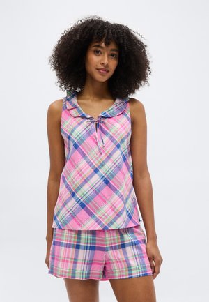 Kobieta o kręconych włosach, ubrana w różowo-niebiesko-zieloną kraciastą bezrękawnikową bluzkę z kołnierzykiem oraz dopasowane szorty, stojąca na białym tle.