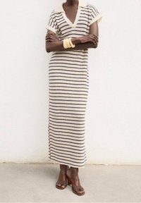 Personne portant une robe longue rayée crème et marine à manches courtes, des bracelets jaunes et des sandales à talons marron, debout les bras croisés.