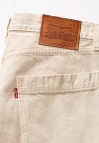 Bež traperice s teksturiranim kožnim oznakom na kojoj piše "Levi Strauss & Co." i crvenom trakicom sa strane, s klasičnim stražnjim džepovima.