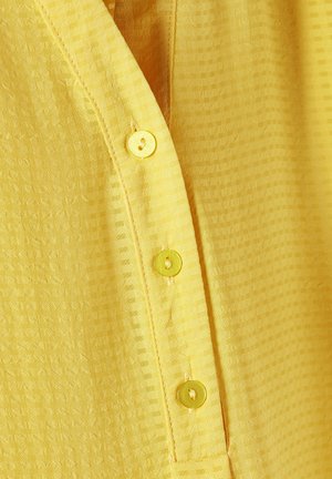 Gul tekstureret skjorte med en stolpe med fire runde, gule knapper og lodrette striber på stoffet.