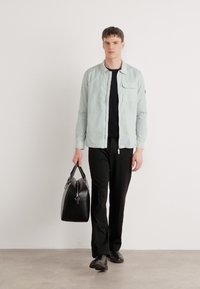 Világoskék csíkozott zakó elülső zsebbel, fekete ing, fekete nadrág és fekete bőr táska; a modell egy semleges háttér előtt sétál.