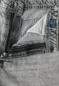 Gros plan d’un jean en denim gris non fermé avec fermeture éclair visible, bouton, couture et doublure blanche de la poche intérieure.