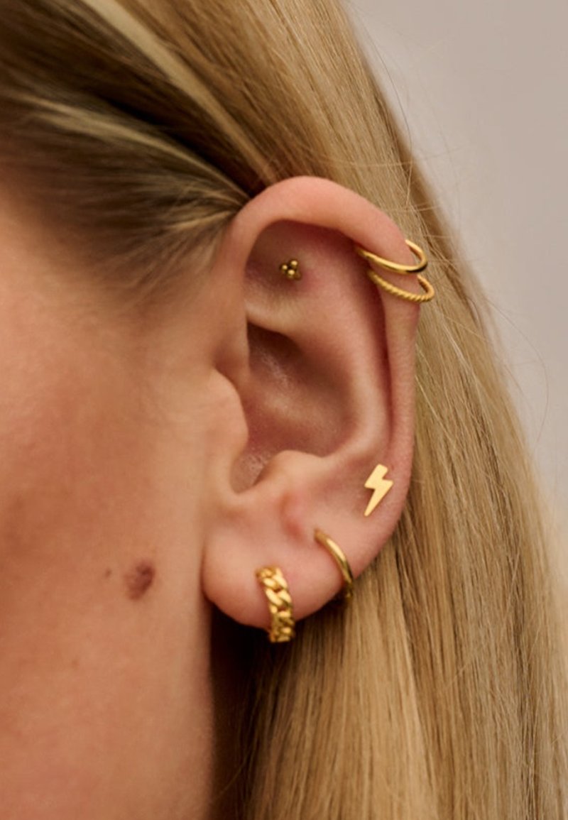 Orecchini d'oro in vari design: due cerchi di filo, un fulmine, una fascia intrecciata e un chiodo con tre piccole sfere. L'orecchio è parzialmente visibile.