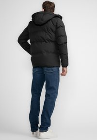 Giacca a vento nera con cappuccio, design trapuntato e polsini elastici, abbinata a jeans blu scuro e scarpe bianche. Vista posteriore.
