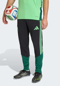 Črne športne hlače z zelenimi poudarki in logotipom Adidas, kombinirane s črnimi kopačkami. V levi roki je večbarvna nogometna žoga.