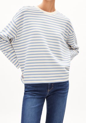 Person iført en løstsiddende langærmet top med lyseblå og hvide vandrette striber og blå cowboybukser, hænderne på hoften.