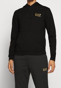 Černá mikina s klokankou, která má na hrudi zlaté logo EA7 a dekorativní design na rukávech. Měkký bavlněný materiál.